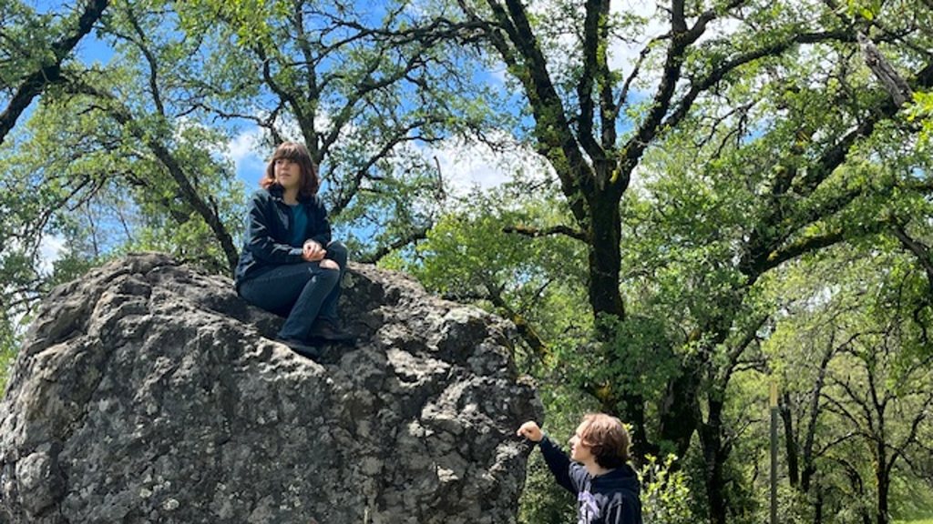 A young actor sits alone on the great rock beneath the oak while another climbs toward the top.