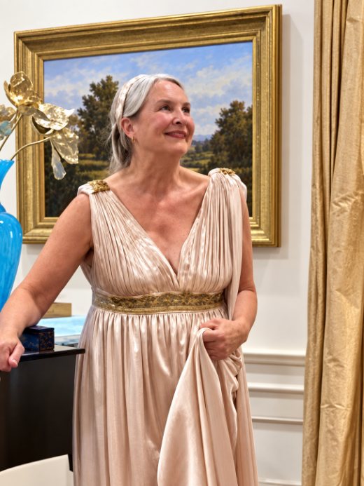 Mari Reeves in a pleated blush silk gown with gold shoulder clasps and an embroidered gold belt, standing before a gilt-framed landscape painting.